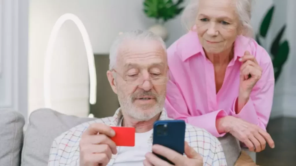 idoso com cartão e celular nas mãos e senhora por tras observando